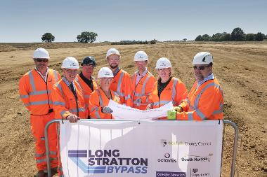 Group of people in orange high-visibility clothing standing on site, behind a banner listing the Long Stratton Bypass funders and construction company.