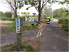 View of signage for the Marriott's Way path, with the level crossing gates marking the start of the Marriott's Way route at Barn Road in the background and a mobility scooter using the path.