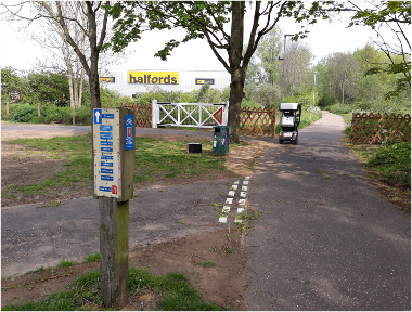 View of signage for the Marriott's Way path, with the level crossing gates marking the start of the Marriott's Way route at Barn Road in the background and a mobility scooter using the path.