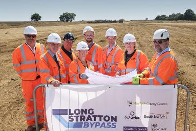 Group of people in orange high-visibility clothing standing on site, behind a banner listing the Long Stratton Bypass funders and construction company.