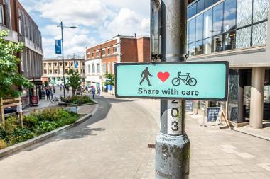 A photograph of a 'Share with Care' sign - featuring a human figure, a heart symbol, and a bicycle - mounted on a lamppost along Westlegate.