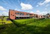 An image of the outside of Cringleford Prep Primary School. The image shows a two storey red and green brick building, with an outside shelter and green space alongside the car park.